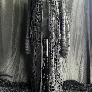 Può includere: Un lungo cardigan all'uncinetto grigio, aperto sul davanti, con un esclusivo design traforato. Il capo fatto a mano presenta maniche lunghe e orlo a terra. Il cardigan è esposto su un manichino su sfondo scuro.