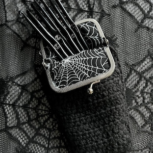 May include: A black crocheted pouch with a zip closure, containing a set of black crochet hooks. The pouch is lined with a black and white spiderweb pattern fabric.