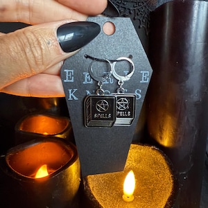 May include: Silver hoop earrings with black enamel book charms. Each charm features the word "SPELLS" and a pentagram. The earrings are displayed on a black tag with the letters "E E K N" visible. The background includes lit candles.