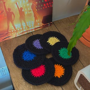 Op de afbeelding: Set van zes gehaakte onderzetters in de vorm van een plaat. Elke onderzetter is zwart met een kleurrijk midden in tinten van paars, geel, blauw, groen, rood en oranje.