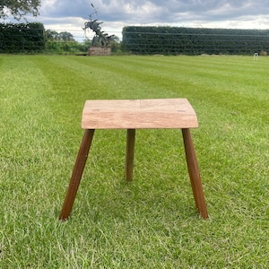 May include: A small, handcrafted wooden stool with a square seat and three angled legs. The seat is light brown, and the legs are a darker brown. The stool is set on a green lawn, suggesting outdoor use. The design is simple and rustic.