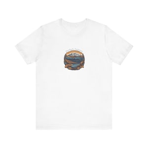 May include: White t-shirt featuring a circular graphic with a landscape design. The graphic depicts a mountain range, a body of water, and the text "Deadloch Tasmania" in a semi-circle above the landscape. The design is in shades of brown, blue, and orange.
