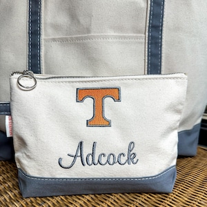 May include: Cream-colored canvas tote bag and matching zippered pouch. The pouch features an orange and white embroidered "T" logo and the name "Adcock" in script. Both items have gray accents and a silver zipper.
