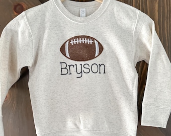 May include: A heathered oatmeal-colored long-sleeve sweatshirt featuring an embroidered brown football with white stitching details centered on the chest. Below the football, the name Bryson is embroidered in a black, simple sans-serif font.