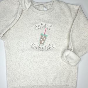 May include: A light gray sweatshirt with the text "Cutest Coffee Date" embroidered on the front. The text is in black with a small cartoon image of a coffee cup with ice cubes and a red straw.