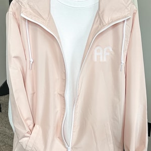 May include: A light pink windbreaker jacket with a white zipper and white drawstring hood. The jacket has a white embroidered "AF" on the front.
