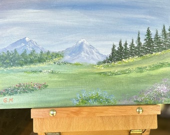 May include: A painting on a wooden easel depicts a landscape with two snow-capped mountains in the distance, a field of green grass with wildflowers, and a row of dark green trees in the foreground. The sky is a light blue with white clouds. The painting is signed "G.M."