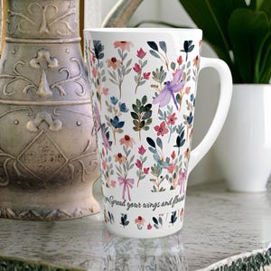 May include: A white ceramic latte mug featuring a watercolor pattern of colorful wildflowers, green leaves, and a purple dragonfly. The text "Spread your wings and flourish" is printed in black script near the base of the mug.