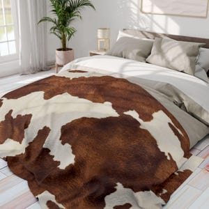 May include: A brown and white cowhide-patterned blanket draped over a bed. The blanket has a soft, textured appearance and is made of a faux fur material. The bed has neutral-colored bedding and pillows.