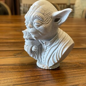 May include: A light gray bust of Yoda from Star Wars, with detailed facial features and large ears. The sculpture depicts Yoda holding Grogu. The bust is made of a speckled material and sits on a wooden surface.