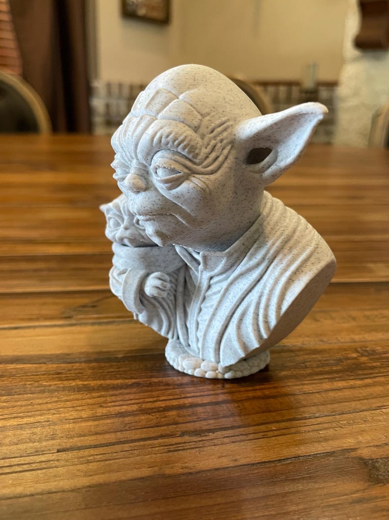 May include: A light gray bust of Yoda from Star Wars, with detailed facial features and large ears. The sculpture depicts Yoda holding Grogu. The bust is made of a speckled material and sits on a wooden surface.