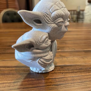 May include: A light gray sculpture of Yoda holding Grogu. The Yoda figure has large ears and detailed facial features. Grogu is cradled in Yoda's arms. The sculpture is on a wooden surface.