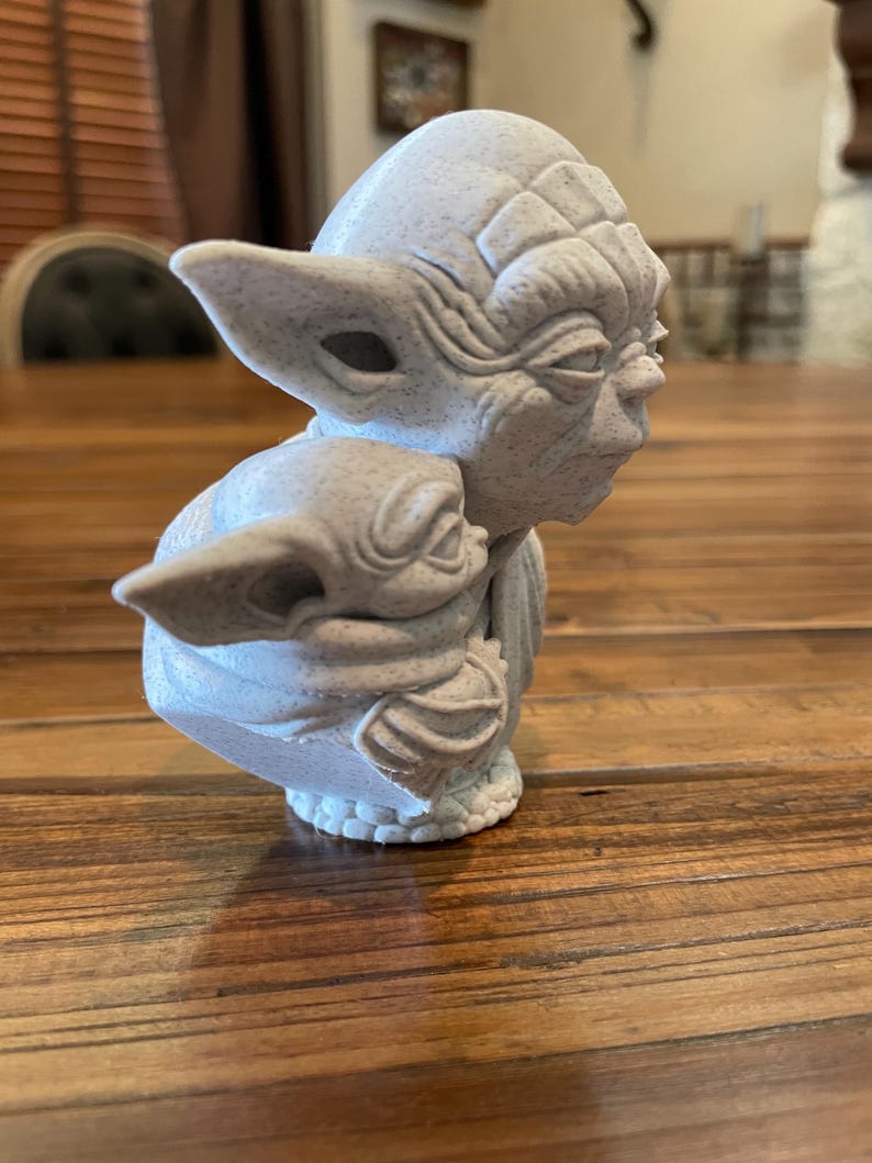 May include: A light gray sculpture of Yoda holding Grogu. The Yoda figure has large ears and detailed facial features. Grogu is cradled in Yoda's arms. The sculpture is on a wooden surface.