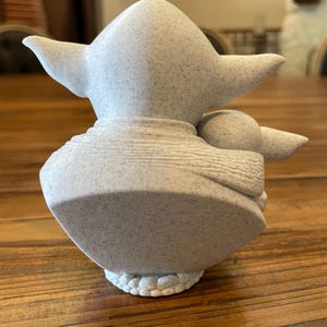 May include: A light gray figurine of a character with large ears and a small body, holding a smaller figure. The figurine has a speckled texture and sits on a textured base. The overall design is reminiscent of a popular science fiction character.