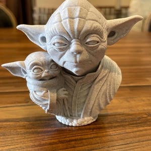 May include: A light gray Yoda statue holding a smaller Baby Yoda figure. The statue has detailed facial features and is set on a wooden surface. The sculpture is a collectible item.