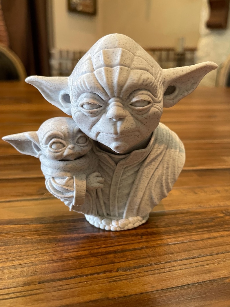 May include: A light gray Yoda statue holding a smaller Baby Yoda figure. The statue has detailed facial features and is set on a wooden surface. The sculpture is a collectible item.