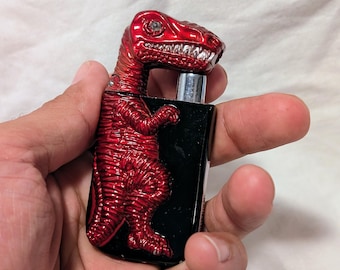 May include: A red dinosaur-shaped lighter with a black base. The lighter is metallic with a textured finish, and the dinosaur's head is at the top. The lighter is held in a hand against a white background.