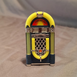 May include: A miniature jukebox with a yellow arch and accents, featuring the text "LIGHT MY FIRE". The jukebox has a dark blue body with silver and yellow details, and a diamond-shaped grill.