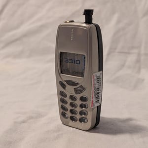 Puede incluir: Un teléfono móvil vintage plateado y negro. La pantalla muestra "3310". El teléfono tiene un teclado con varios botones y una antena. Una pegatina está pegada en el lateral. Diseño clásico.