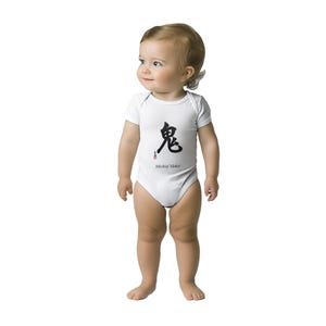May include: A white short-sleeved baby bodysuit featuring a black Japanese kanji character for oni, a small red stamp-like mark below it, and the text "Mischief Maker" printed in black lettering on the front centre of the garment.