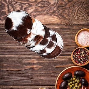 May include: A set of six brown and white cowhide coasters. The coasters are stacked on top of each other on a wooden surface.