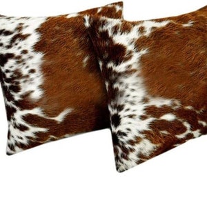 May include: Two brown and white cowhide throw pillows. The pillows have a soft, fuzzy texture and are perfect for adding a touch of rustic charm to any room.