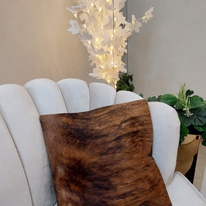 Op de afbeelding: Een witte bank met een bruin en zwart koeienhuid kussen. De bank staat in een kamer met een witte boom met lampjes en groene planten.