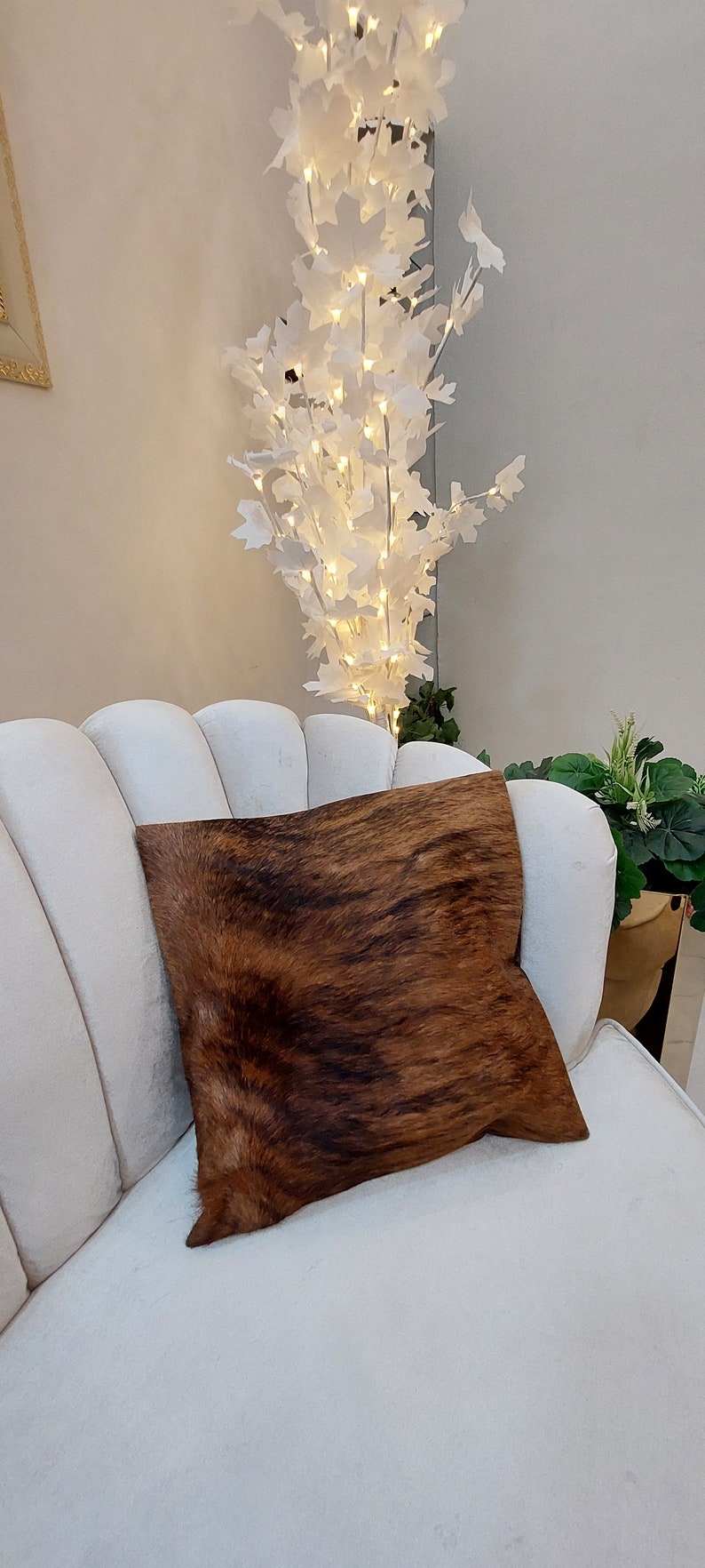 Op de afbeelding: Een witte bank met een bruin en zwart koeienhuid kussen. De bank staat in een kamer met een witte boom met lampjes en groene planten.