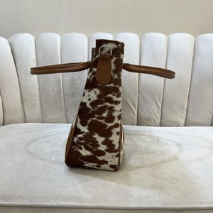 May include: A cowhide tote bag with brown and white spotted pattern. The bag has brown leather handles and trim. The bag is sitting on a white surface.