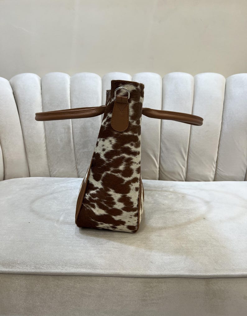 May include: A cowhide tote bag with brown and white spotted pattern. The bag has brown leather handles and trim. The bag is sitting on a white surface.