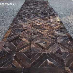 K&ouml;nnte beinhalten: Ein brauner Leder-Patchwork-Teppich mit geometrischem Muster. Der Teppich hat einen dunkelbraunen Rand und besteht aus verschiedenen Lederst&uuml;cken, die zusammengen&auml;ht sind.