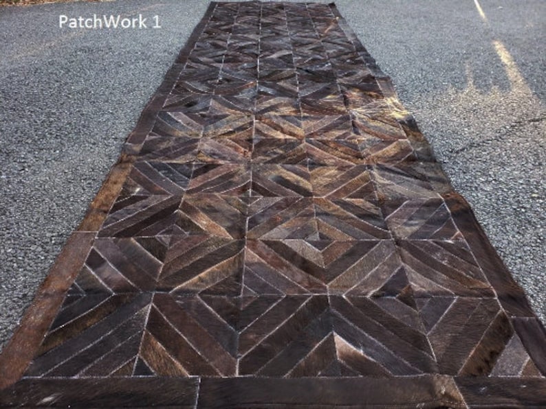K&ouml;nnte beinhalten: Ein brauner Leder-Patchwork-Teppich mit geometrischem Muster. Der Teppich hat einen dunkelbraunen Rand und besteht aus verschiedenen Lederst&uuml;cken, die zusammengen&auml;ht sind.