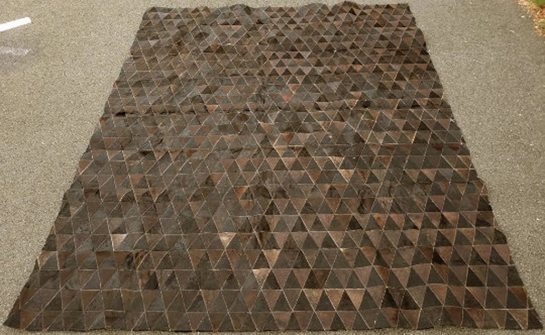 K&ouml;nnte beinhalten: Ein brauner und schwarzer geometrischer Teppich aus Lederdreiecken. Der Teppich hat einen dezenten Glanz und liegt flach auf einer grauen Oberfl&auml;che.
