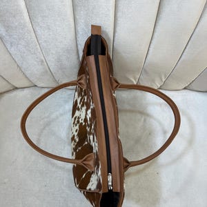 May include: A brown and white cowhide tote bag with brown leather trim and handles. The bag has a black zipper closure and a silver-colored zipper pull. The bag is designed with a spacious interior and is suitable for everyday use.