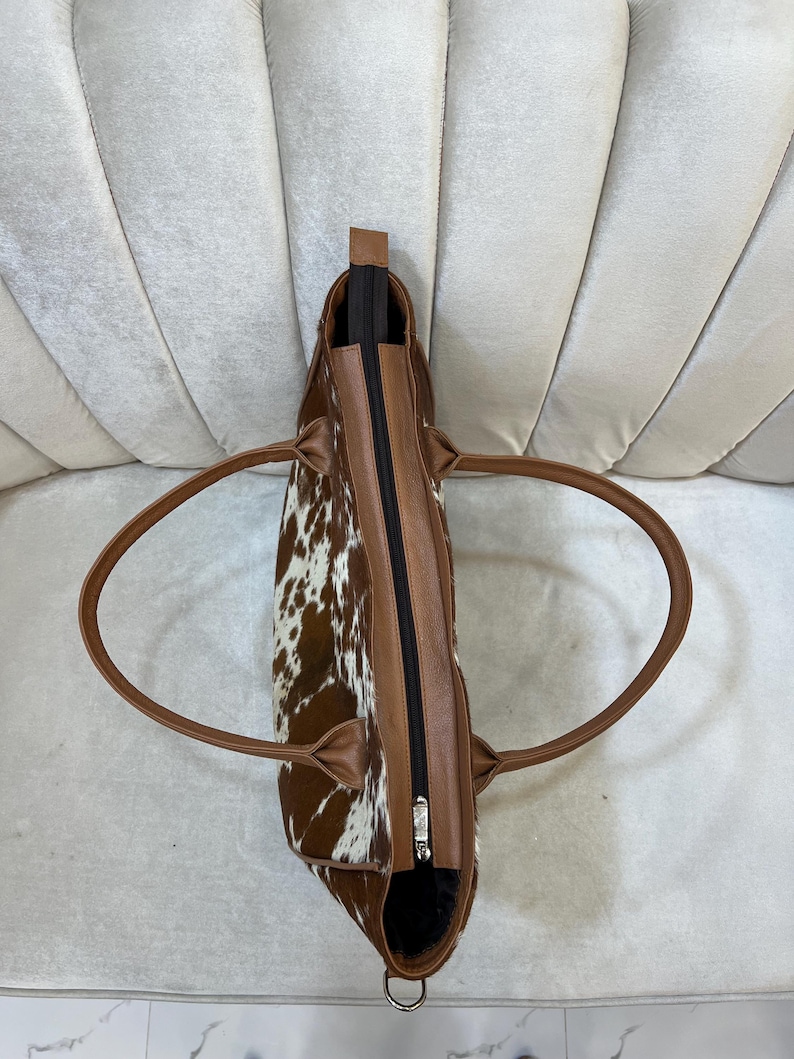 May include: A brown and white cowhide tote bag with brown leather trim and handles. The bag has a black zipper closure and a silver-colored zipper pull. The bag is designed with a spacious interior and is suitable for everyday use.