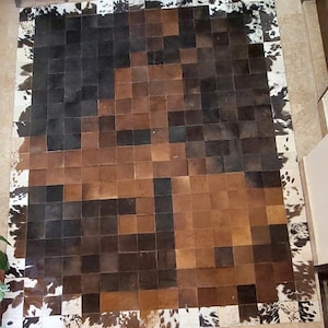 May include: A large, rectangular cowhide rug with a patchwork design. The rug features squares of brown, black, and tan leather, with a border of white and brown cowhide. The rug is a decorative floor covering.