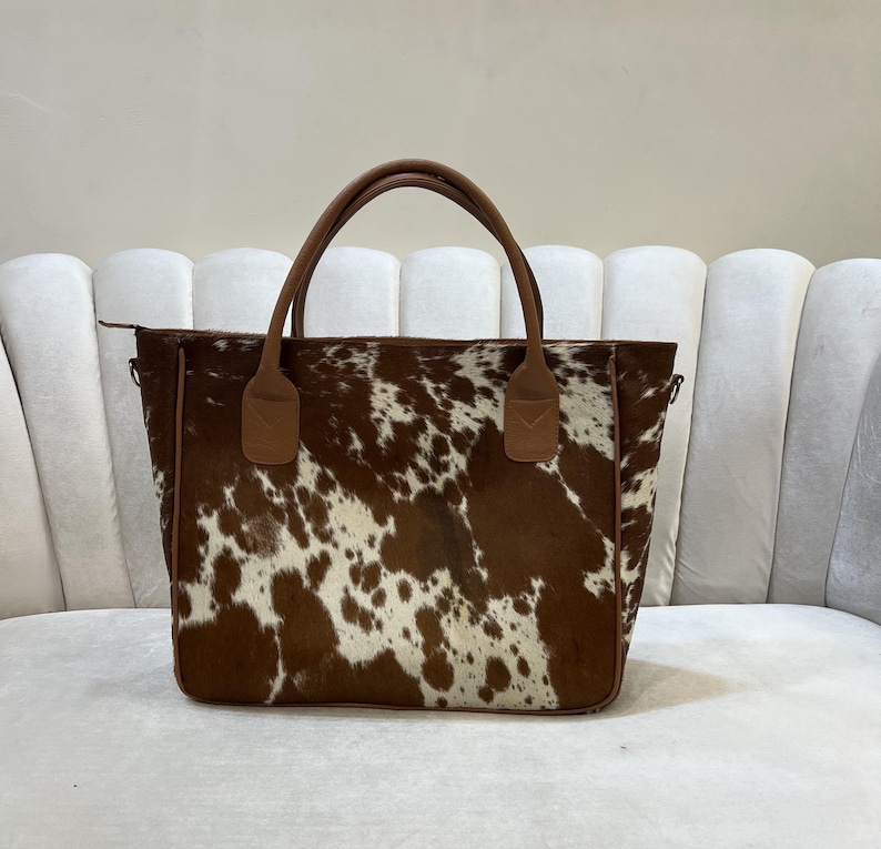 May include: A brown and white cowhide tote bag with brown leather handles and trim. The bag has a rectangular shape and a zipper closure. The handles are attached with brown leather accents. The bag is sitting on a white surface.