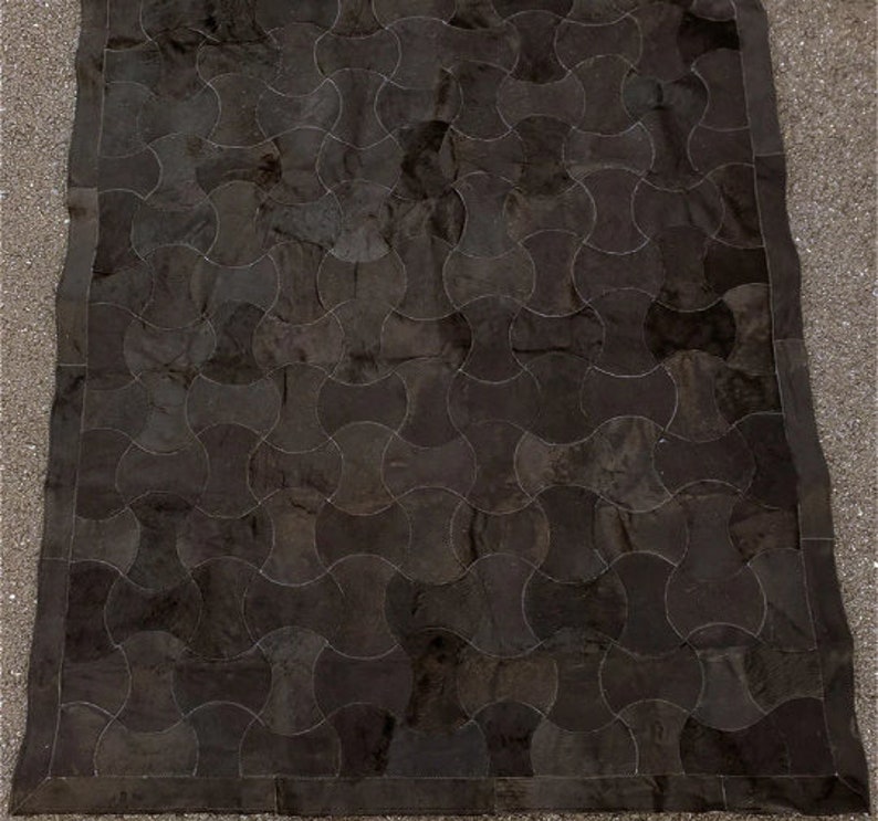 K&ouml;nnte beinhalten: Ein schwarzer Lederteppich mit einem geometrischen Muster aus &uuml;berlappenden Kreisen. Der Teppich hat einen dunkelbraunen Rand.