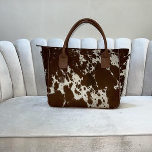 May include: A brown and white cowhide tote bag with brown leather handles. The bag has a rectangular shape and a zipper closure. The bag is sitting on a white couch.