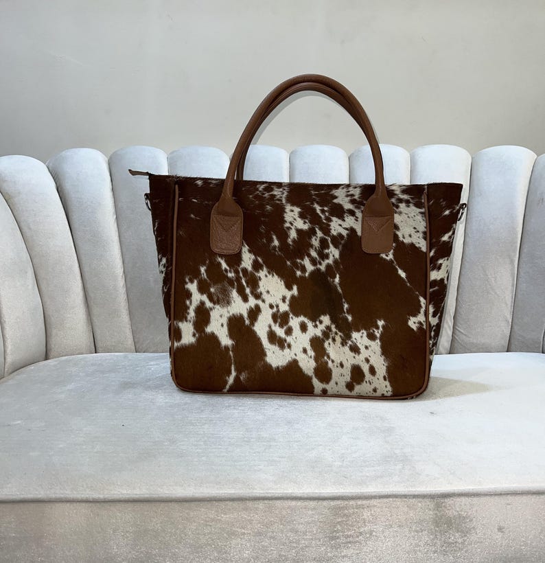 May include: A brown and white cowhide tote bag with brown leather handles. The bag has a rectangular shape and a zipper closure. The bag is sitting on a white couch.