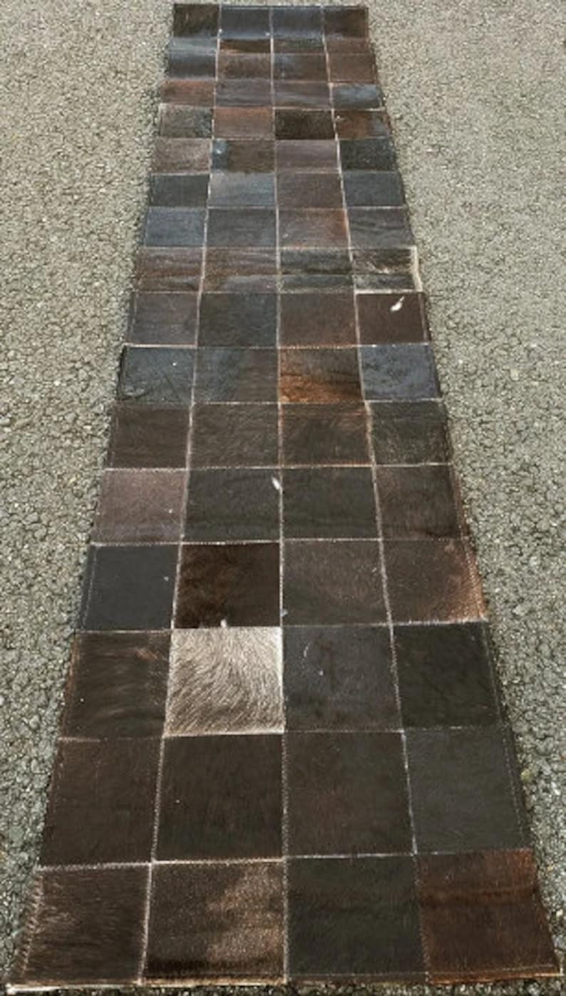 K&ouml;nnte beinhalten: Ein langer, rechteckiger Teppich aus braunen und wei&szlig;en Kuhfell-Flicken. Der Teppich besteht aus vielen Quadraten, die jeweils ein anderes Muster und eine andere Textur haben.