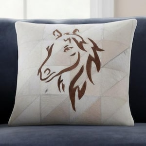 May include: Square decorative pillow with a patchwork design in neutral tones. A brown horse head silhouette is centered on the pillow. The pillow is placed on a dark blue sofa.