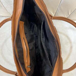 May include: Open view of a brown leather tote bag with a zipper closure. The bag has a light brown interior with multiple pockets and a dark brown lining. The exterior features a textured, light brown and white pattern.