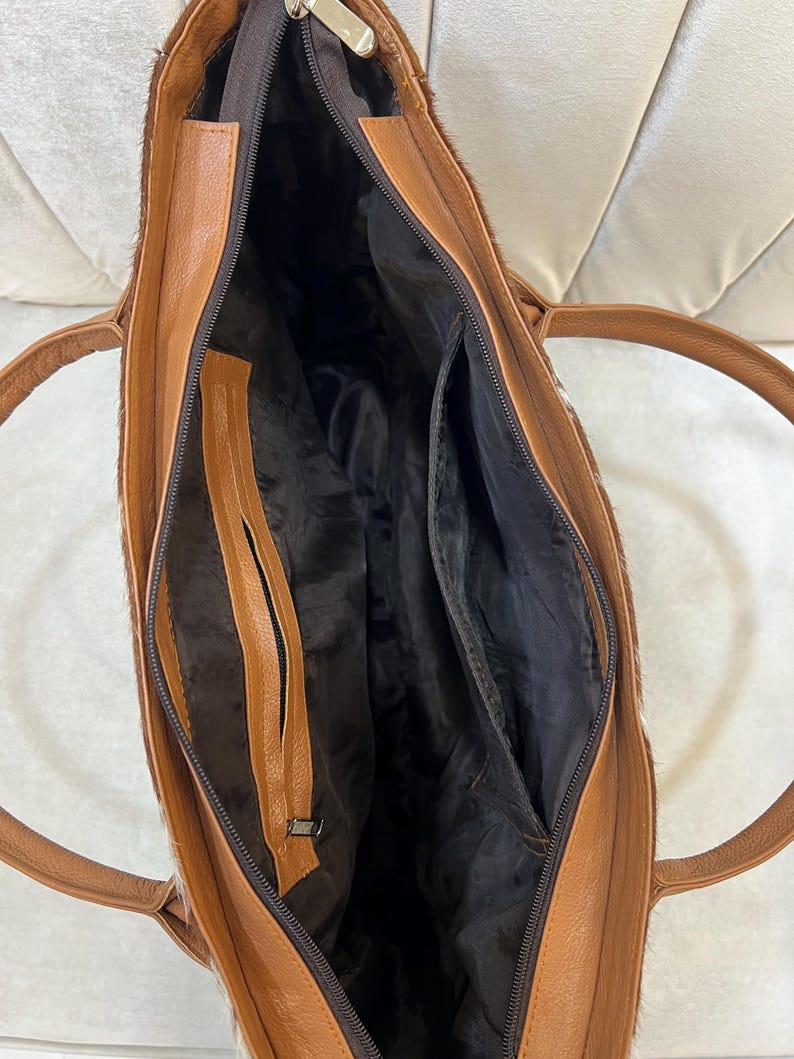 May include: Open view of a brown leather tote bag with a zipper closure. The bag has a light brown interior with multiple pockets and a dark brown lining. The exterior features a textured, light brown and white pattern.