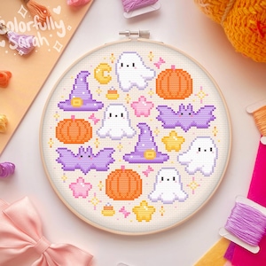 May include: A white embroidery hoop with a colorful cross-stitch design featuring ghosts, bats, pumpkins, stars, and witch hats. The design is surrounded by a light brown wooden hoop.