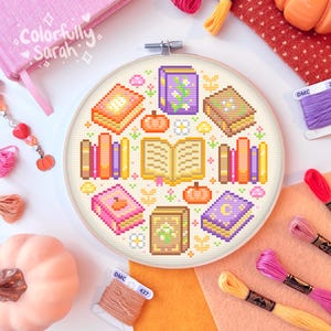 Puede incluir: Un bastidor de bordado de punto de cruz con un diseño de libros, calabazas y elementos florales. La paleta de colores incluye naranja, rosa, morado y amarillo. El texto "Colorfully Sarah" es visible en un cuaderno rosa. También hay hilo de bordar y una calabaza.