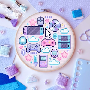 May include: A cross-stitch hoop featuring a pastel-colored video game design. The design includes game controllers, a mouse, a CD, and a game console. The hoop is surrounded by spools of thread, felt, and other craft supplies. The text "Colorful Sarah" is visible.