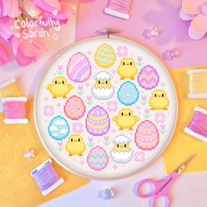 Könnte beinhalten: Ein Kreuzstichmuster mit einem farbenfrohen Osterdesign mit Küken und Eiern. Das Muster ist in einem weißen Stickrahmen mit Holzrahmen dargestellt.