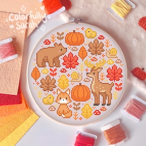 May include: A round cross-stitch embroidery hoop featuring a colourful autumn scene. The design includes a fox, a bear, a deer, pumpkins, leaves and mushrooms. The hoop is made of wood and has a white background.