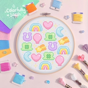 Könnte beinhalten: Ein weißer Stickrahmen mit hellblauem Hintergrund und einem bunten Kreuzstich-Design. Das Design zeigt Regenbögen, Herzen, Kleeblätter, Sterne, Hufeisen und Monde. Der Text "Colorfully Sarah" steht oben links.